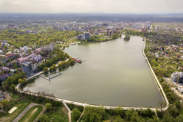 Aerial view of city lake among green trees and town buildings in recreation park zone. Drone