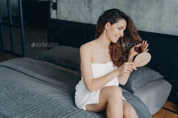 Gorgeous european woman combs her long silky hair in bedroom. Femininity and beauty concept.
