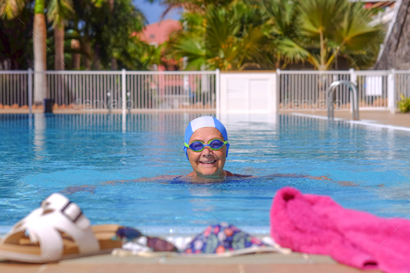 Smiling senior woman swims in the outdoor swimming pool wearing swimming cap and goggles