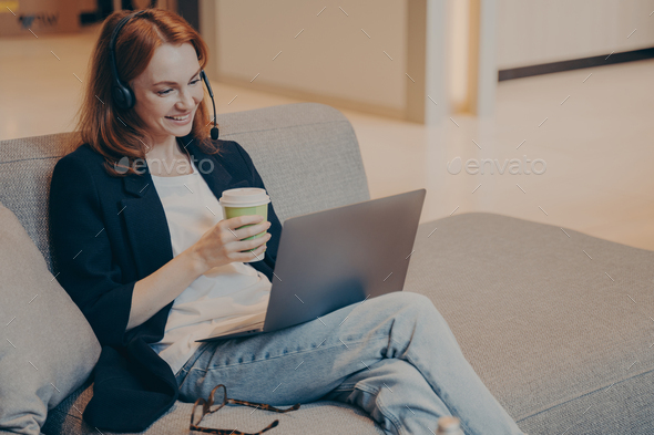 Smiling young female call center agent in wireless headset and laptop sitting on sofa in cafe