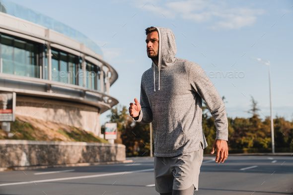 Young male jogger wearing grey hoodie running across the street in urban background