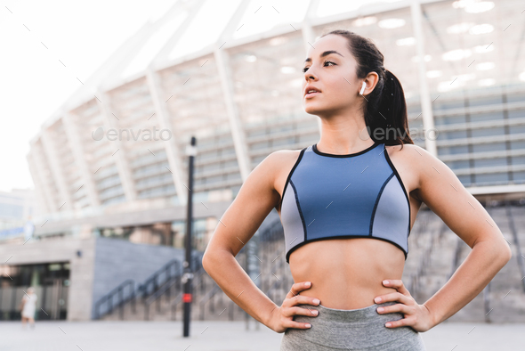 Confident fit caucasian female runner standing against big modern stadium in sporty top with earbuds