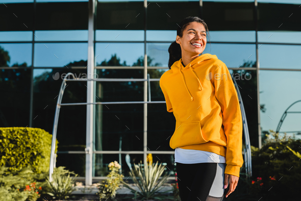 Smiling young fit afro girl wearing bright yellow hoodie resting after jogging in urban area