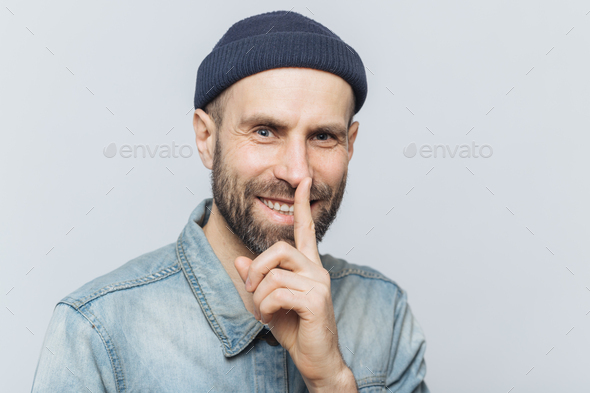 Cheerful attractive male with blue eyes shows silence sign, has satisfied expression.