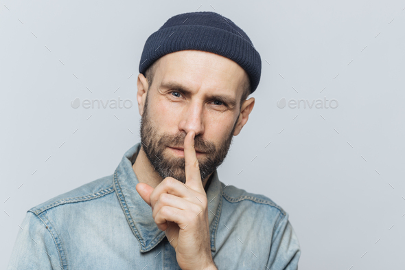 Handsome man with thick beard and mustache, has secret look, demonstrates silence sign.