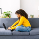 Smiling young woman with curly hair using laptop while relaxing on sofa at home - PhotoDune Item for Sale