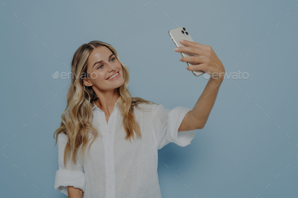 Beautiful young female with long blonde dyed hair holding mobile phone and posing for selfie