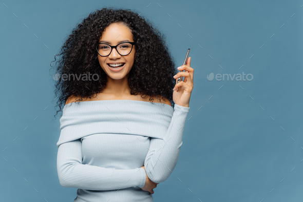 Waist up shot of mirthful Afro American woman holds smart phone, waits for call