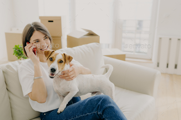 Cheerful woman poses on comfortable sofa, speaks with friend via smartphone