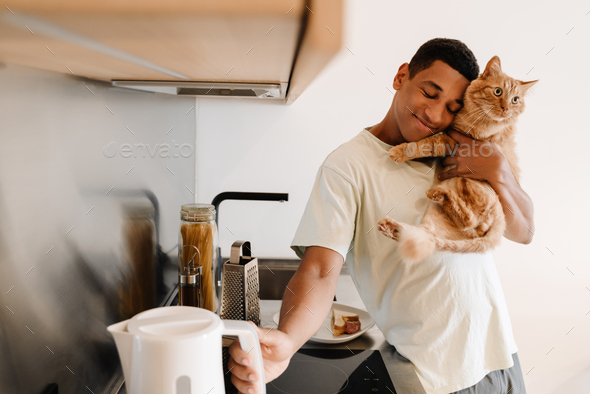 Black man wearing t-shirt holding his cat while making breakfast