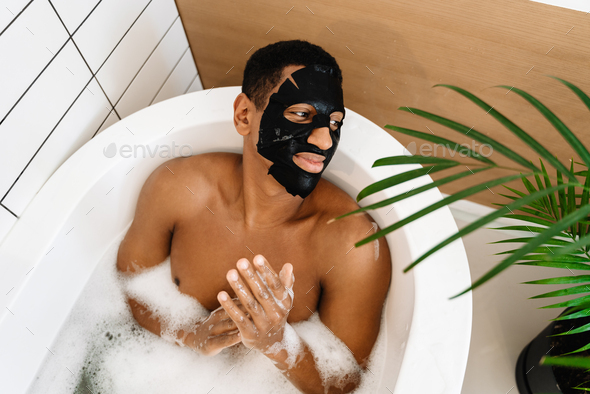 Black naked man using cosmetic mask taking bath with foam