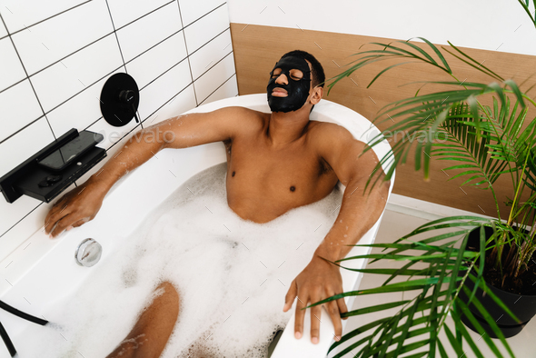 Black naked man using cosmetic mask taking bath with foam