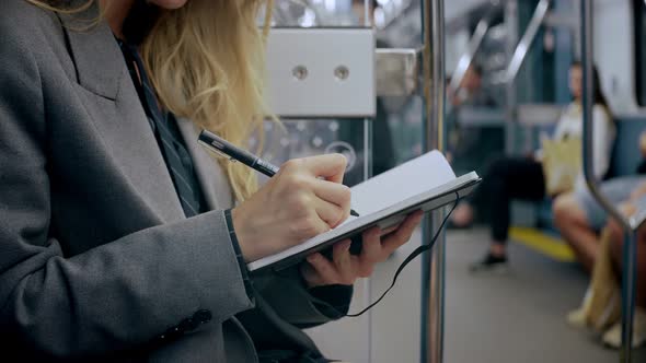 Blonde Business Girl Writing in Black Notebook Sitting in Modern Metro Train