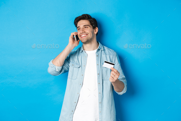 Smiling man call bank support, talking on mobile phone and holding credit card, standing against