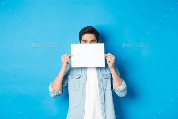 Sneeky handsome guy hiding face behind blank piece of paper for your logo, making announcement or