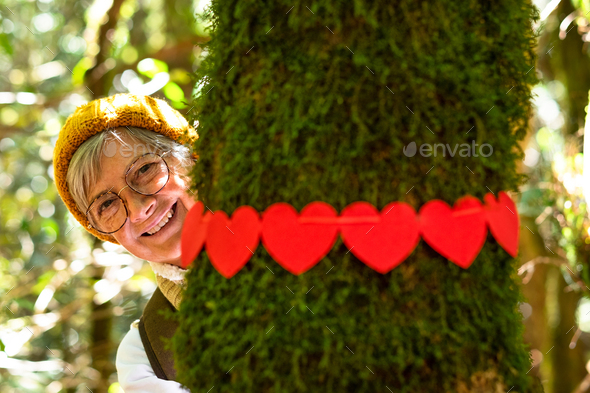 Smiling elderly woman hugging a moss covered tree trunk in the woods with a wreath of red hearts