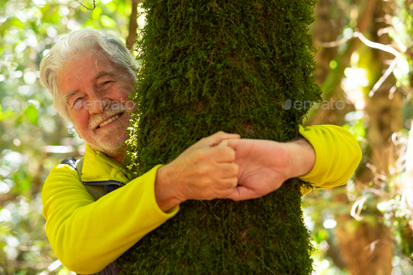 Smiling partially hidden elderly man hugging a moss covered tree trunk in the woods with his arms