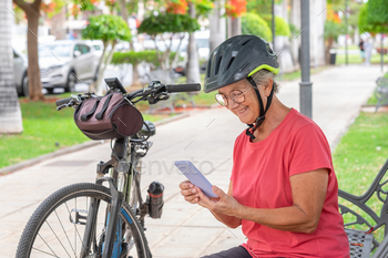 Active cyclist elderly woman sitting on a bench in public park reading message on mobile phone