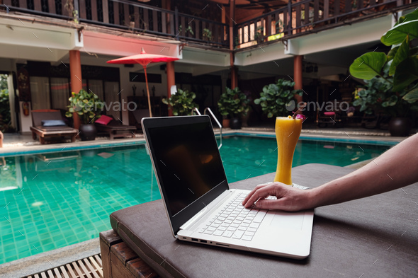 travel blogger sitting at swiming pool writing article on white laptop.
