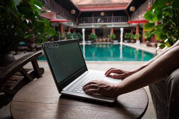 travel blogger sitting at swiming pool writing article on white laptop.