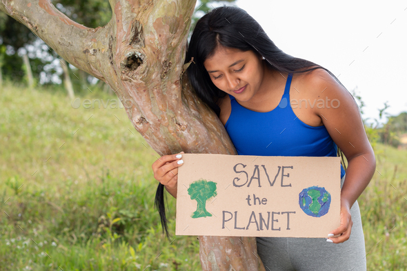 A brunette woman holds a sign calling to save the planet as she reads it. Latina girl protects tree.