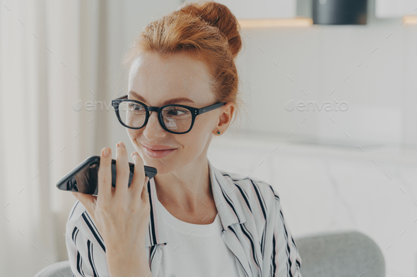 Horizontal shot of ginger woman uses voice assistant on smartphone makes voice call