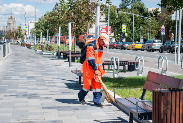 Municipal worker in orange uniform collecting garbage from sidewalk.
