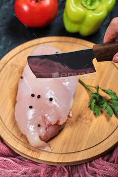 Hands cutting piece of raw chicken fillet on wooden plate with fresh vegetables and tablecloth