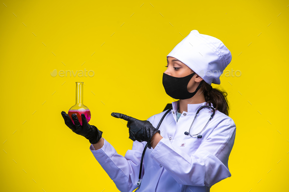 Young nurse in face and hand mask holds chemical flask and points at it with finger