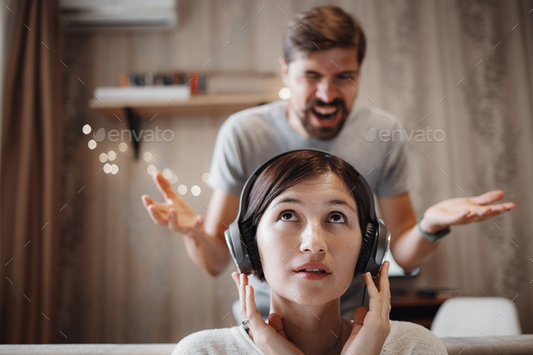 angry husband shouting at wife sitting on sofa and covering ears with headphones