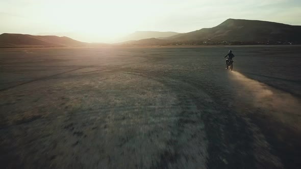 Drone View of Man on Motorbike Rides on a Dry Lake with Dust and Beautiful Hills on Background in