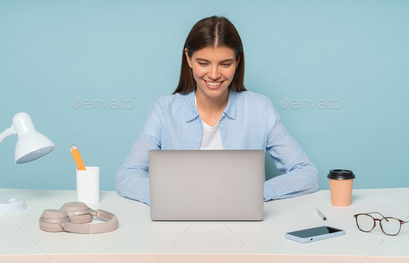 Cute secretary in shirt sitting at desk working on laptop over blue studio background