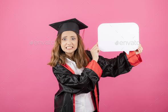 Annoyed young girl holding white idea board and wearing graduation gown on pink background