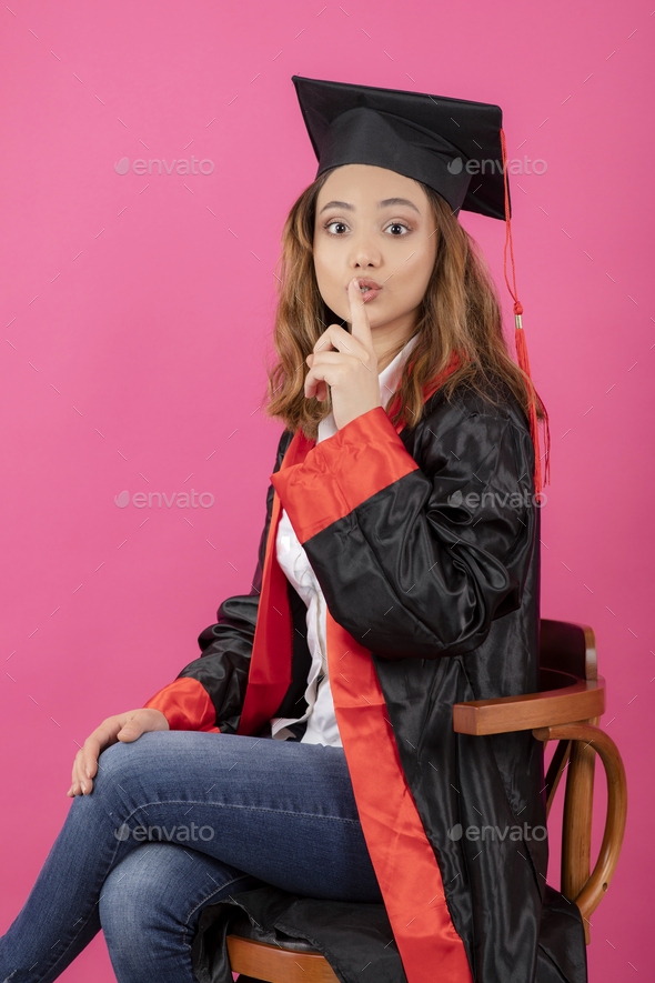 Portrait of female student wearing graduation gown and gesture silent on pink background