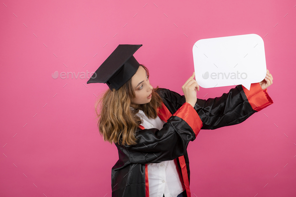 Female student wearing graduation gown and holding white idea board on pink background