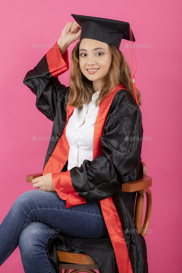 Young female student wearing graduation gown and sitting on a chair on pink background