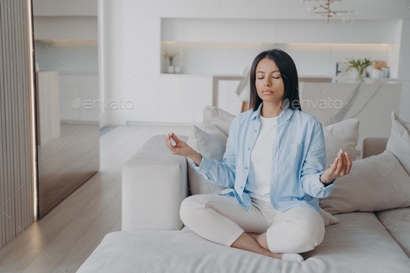 Woman practice yoga, meditating, breathing, relaxing in lotus pose on couch at home. Stress relief