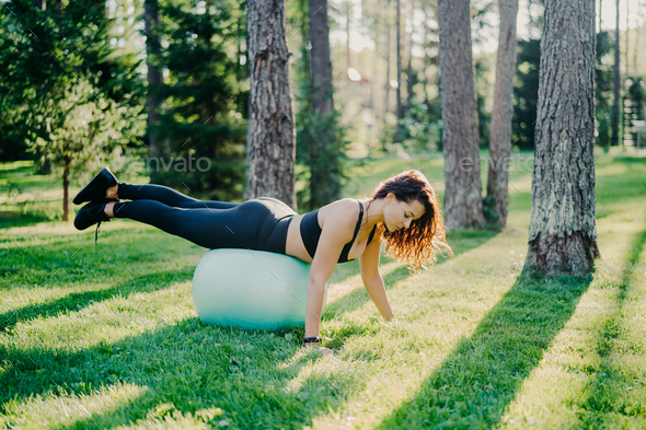 Outdoor shot of sporty young woman leans at fitness ball keeps balance
