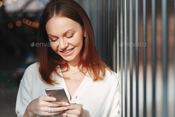 Pleased woman focused into screen of cell phone, checks email box, dressed in white clothes