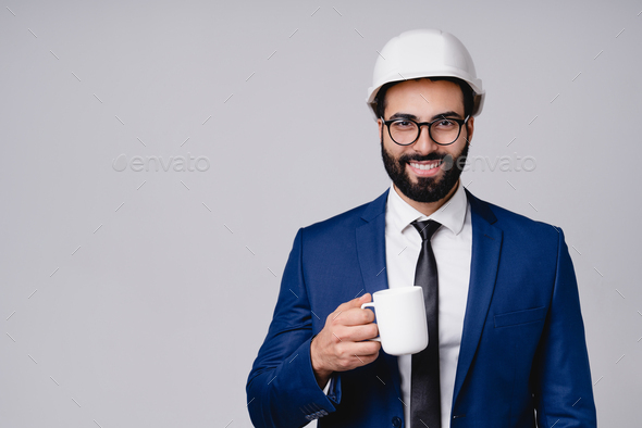 Busy successful young moroccan engineer in hardhat with cup of coffee isolated over grey background