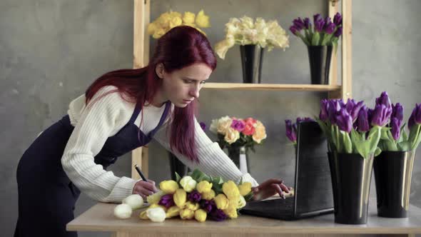 Florist in an Apron Working in Her Own Flower Shop Using a Laptop and Writing Down an Order for a alt