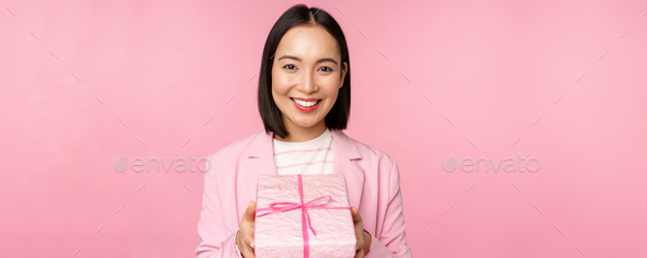 Smiling asian businesswoman in suit, giving you gift in wrapped box, standing over pink background