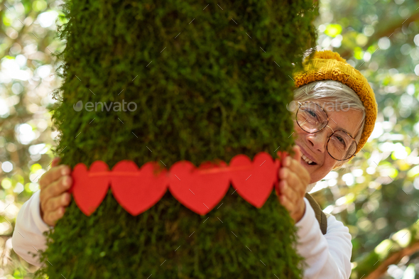 Smiling partially hidden elderly woman hugging a moss covered tree trunk in the woods