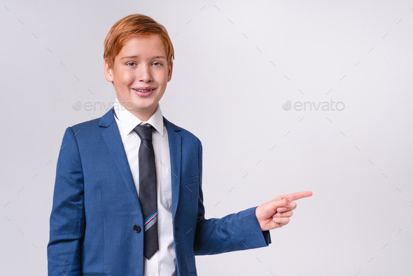Redhead preteen boy with braces in formal attire pointing at copy space isolated over grey