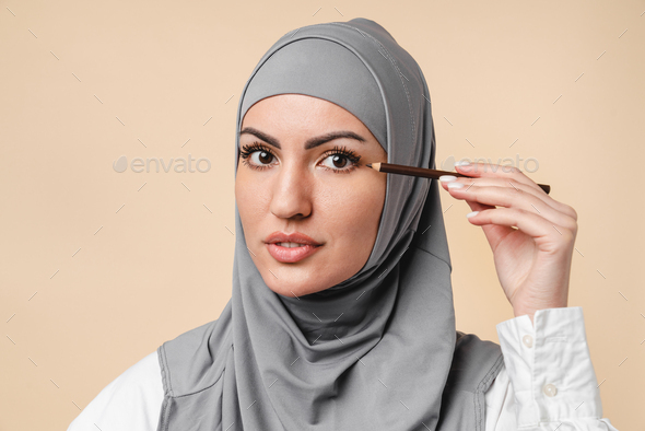 Pretty young arabian muslim islamic woman wearing grey hijab applying makeup with eye pencil