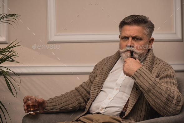 handsome senior man smoking pipe in armchair and looking at camera
