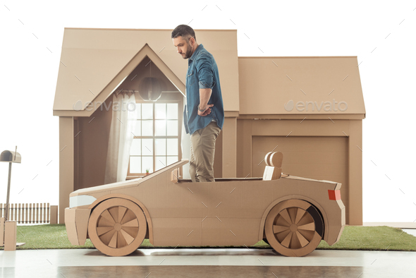 side view of man in cardboard car in front of cardboard house isolated on white