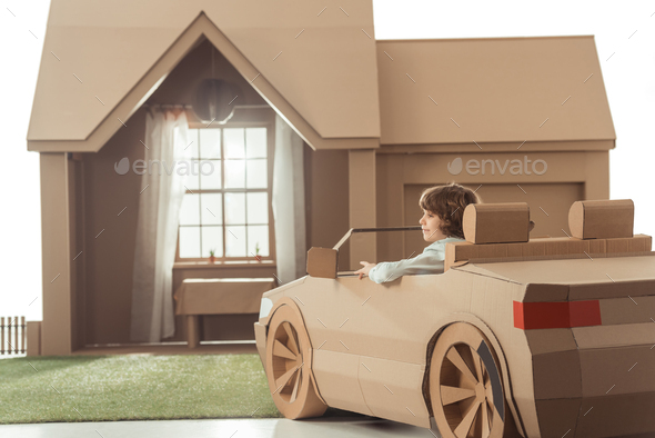 little kid sitting in cardboard car in front of cardboard house isolated on white