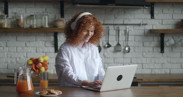 Redhead Smiling Young Woman with Curly Hair Putting on Headphones While Working at Laptop Computer alt