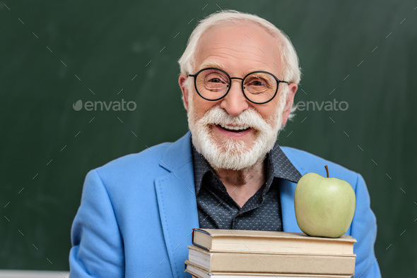 smiling grey hair professor holding stack of books with apple on top